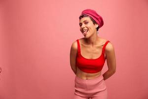 Young athletic woman with a short haircut and purple hair in a red top and a pink hat with an athletic figure smiles and grimaces looking at the camera on a pink background photo