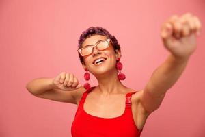 A young athletic woman with a short haircut and purple hair in a red top in sunglasses with an athletic figure smiles with her arms outstretched in different directions dancing on a pink background photo