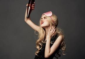 Drunk woman with a bottle of alcohol in her hand on a gray background and a mask for sleeping photo