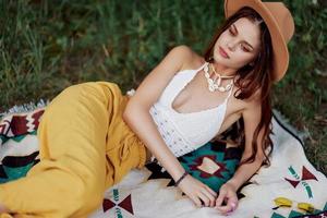 un mujer en Respetuoso del medio ambiente ropa en un hippie Mira es acostado en un de colores tartán sonriente y mirando a un otoño puesta de sol en naturaleza en el parque. estilo de vida en un viaje de armonía con el mundo foto