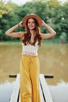 un joven mujer en un hippie Mira y eco ropa viajes al aire libre por el lago vistiendo un sombrero y amarillo pantalones en el otoño foto