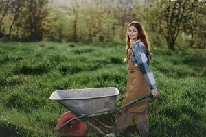 un joven mujer rollos un jardín carro con suelo para plantando en su verde naturaleza jardín y sonrisas foto