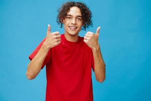 Happy cheerful positive curly tanned Caucasian guy in basic red t-shirt show thumbs up posing isolated on over blue background. Lifestyle and Emotions concept. Good offer with copy space for ad photo