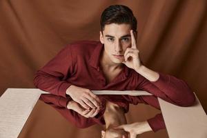 A man on a cloth background with folds sits at a table with a mirror reflection of the model photo