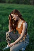 A beautiful redheaded woman sits on the grass in sporty summer clothes and relaxes after a walk in nature and looks out at the sunset. photo