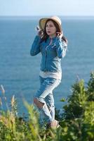 Full length woman in straw hat, denim jacket, jeans and boots standing on seashore in sunny weather photo