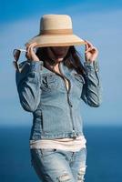 mujer en mezclilla chaqueta y pantalones ocultación con Paja sombrero terminado su cara en antecedentes de cielo y Oceano foto