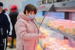An adult woman in a medical mask and glasses examines a label of cheese in a grocery store. photo
