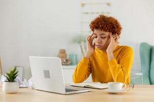 African woman touching temples experiencing stress. Girl at home office tired of working feeling headache sick rubbing temples forehead. Long laptop usage computer vision syndrome. Office syndrome. photo