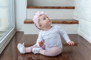 Little crawling baby girl one year old siting on floor in bright light living room near window smiling and laughing photo