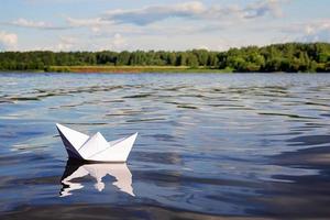 pequeño papel barco flotante en azul calma río agua, verde bosque, azul cielo con ligero verano nubes en el horizonte. libertad, aventura, sueño y viaje concepto foto