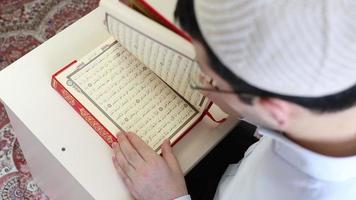 muçulmano adolescente com óculos e religioso chapéu lendo a Alcorão em pé às a mesa, esclarecido imagem do Alcorão video