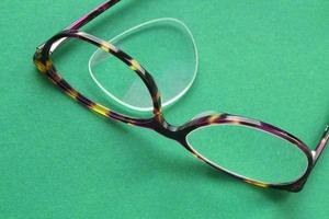 Old broken eyeglasses with damaged lens on green background. Poor eyesight. Reuse and repair concept. Idea of health. Failure optic eyewear. Breakage of vision correction glasses. Close up, flat lay photo