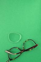 Old broken eyeglasses with damaged lens on green background. Poor eyesight. Reuse and repair concept. Idea of health. Failure optic eyewear. Breakage of vision correction glasses. Close up, flat lay photo