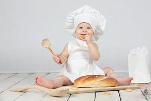 Child cook. Beautiful little girl in cook clothes with a long loaf and a wooden spoon in flour. The child is one year old. Funny baby. Little chef. The concept of baking bakery products. photo