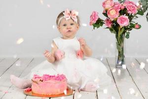 One year old child with a birthday cake. Funny little girl smeared with cream on her birthday. Funny child on holiday. Cute baby girl with pink pastries is sitting on the floor. photo
