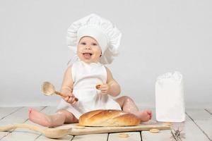 Child cook. Beautiful little girl in cook clothes with long loaf and flour. The child is one year old. Funny baby is laughing. Little chef. The concept of baking bakery products. photo