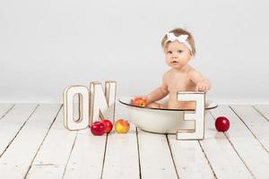 One-year-old child bathes in a basin. Beautiful baby girl with apples. First birthday girl. photo