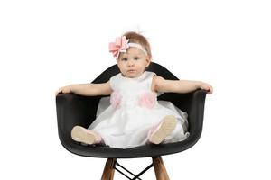 The child is one year old. Beautiful little girl sits on a chair on a white background. Funny child with a bow. Charging baby. photo