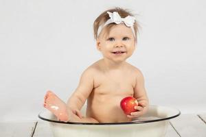 hermosa bebé se baña en un cuenca y come un rojo manzana. pequeño niña en uno año viejo. gracioso niño. foto