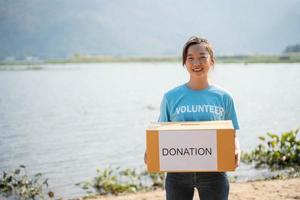 Portrait of young woman wearing volunteer t-shirt,  holding donations box and looking at camera, standing on river background photo