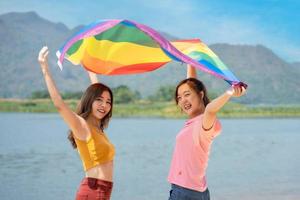 contento lesbiana, hermosa asiático joven dos mujer, niña homosexual, Pareja amor momento gasto bueno hora juntos, levantamiento y ondulación el lgbt bandera en el playa foto