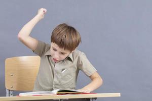 un primario colegio estudiante es haciendo tarea. el niño a el escritorio con el libro de texto es enojado. el chico lo hace no querer a estudiar. un niño de siete años niño va loco para lecciones foto