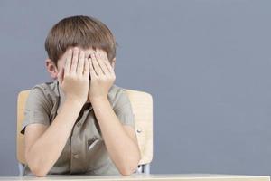 A child sits at a table covering his face with his hands. photo