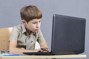 el niño a el ordenador portátil. el chico mira a el monitor pantalla. preescolar a el computadora. júnior estudiante a un mesa con un computadora. foto