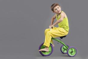 el niño en un triciclo en un gris antecedentes. gracioso pequeño chico es jugando el chico paseos un bicicleta. foto
