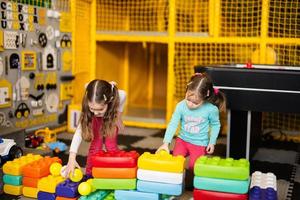 Two sisters playing at kids play center while build with colored plastic blocks. photo