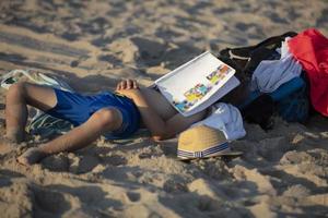 A child sleeps on the beach with his face covered by a magazine. photo