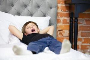 The child yawns while lying on the bed. The boy wants to sleep. Tired elementary school student. photo