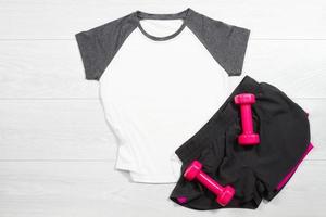 White female sport t shirt mock up flat lay on wooden background. Summer accessories. Sporty trousers, pink female dumbbells. Top front view t-shirt. Mockup tshirt. Template blank copy space photo