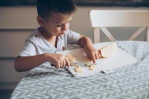 linda chico 4-5 años antiguo con cuchillo corte un queso en el corte tablero en el mesa en el cocina foto