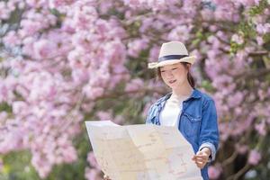 asiático mujer turista participación ciudad mapa mientras caminando en el parque a Cereza florecer árbol durante primavera sakura flor festival concepto foto