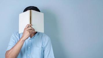 Confident Asian man covering his face with notebook on light blue background photo