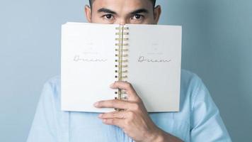 Confident Asian man covering his face with notebook and looking at camera while standing on light blue background photo