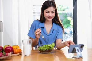 A young woman with a beautiful face in a blue shirt with long hair eating fruit sitting inside the kitchen at home with a laptop and notebook for relaxation, Concept Vacation. photo
