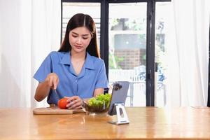 A young woman with a beautiful face in a blue shirt with long hair eating fruit sitting inside the kitchen at home with a laptop and notebook for relaxation, Concept Vacation. photo