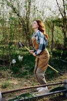 The woman hops amusingly with her rake like a witch on a broomstick in her green garden among the trees and smiles. The concept of everyday lifestyle and joy in the moment photo