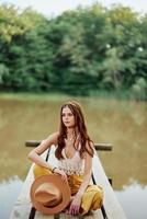 un joven mujer sonriente en un imagen de un hippie y eco-ropa sentado al aire libre en un puente por un lago vistiendo un sombrero y amarillo pantalones en el verano puesta de sol foto