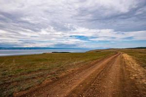 A dirt brown road stretches into the distance along the shore of a lake in cloudy weather. Behind the lake are mountains, the sky is overcast. photo