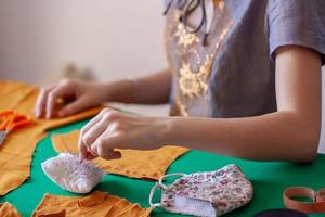 A young girl seamstress takes a needle from a pillow for needles. Orange cloth on a green table. Focus on a pad with needles and fingers. Nearby lies a face mask. Horizontal. photo