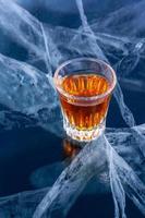 Whiskey in a small glass stands on the transparent ice of Lake Baikal. Deep beautiful cracks in the ice. Focus on a glass of alcohol. Vertical. photo