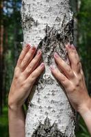 A girl with beautiful nails with two hands hugs a birch in the forest. Face is not visible. The sun is shining. Selective focus on the hand and birch. Ecological concept. photo