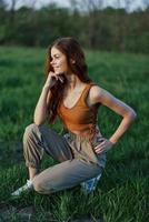 A beautiful redheaded woman sits on the grass in sporty summer clothes and relaxes after a walk in nature and looks out at the sunset. photo