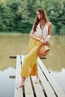 un joven mujer en un hippie Mira y eco-vestido bailando al aire libre por el lago vistiendo un sombrero y amarillo pantalones en el verano puesta de sol foto