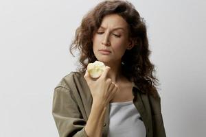 Confused pensive curly beautiful woman in casual khaki green shirt eat apple looks at it with disbelief posing isolated on over white background. Natural Eco-friendly products concept. Copy space photo