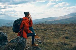 Traveler In a red jacket, hats with a backpack sits on a stone in the mountains in nature photo